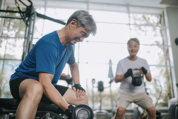 Person helps another use gym equipment, surrounded by weights and machines in a therapy setting.