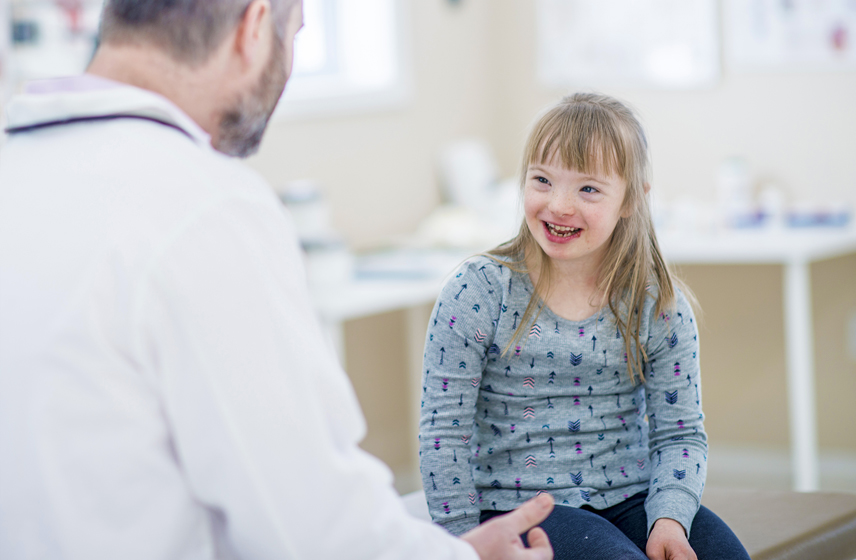 Doctor sitting down, talking with young girl diagnosed with Down Syndrome.