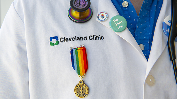 Photo of physician wearing white coat with the Cleveland Clinic logo, a rainbow lapel pin, and a pronoun pin that says he him his