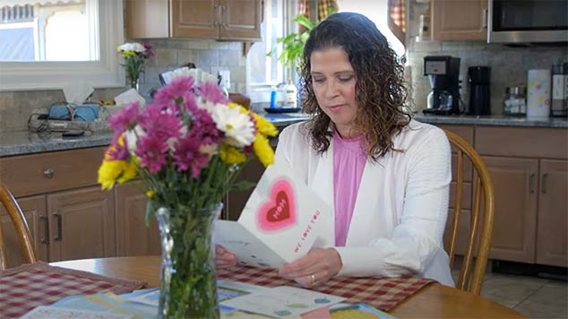 Woman reading a card and sitting at kitchen table with a bouquet of flowers. 