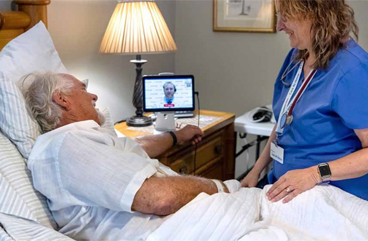 A professional caregiver stands at the bedside of a patient with a mobile device facilitating a virtual checkup