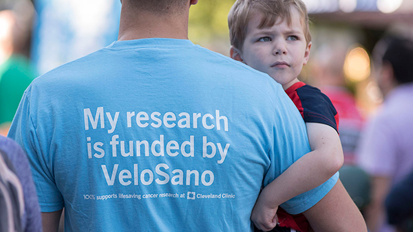 Father and son watching VeloSano riders. Father's shirt says "My research is funded by VeloSano".