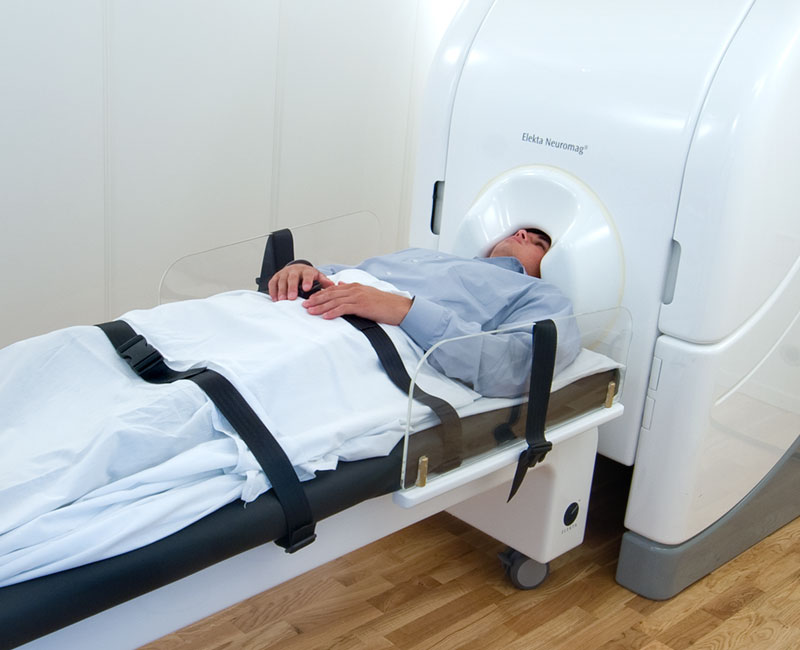 Man sitting slightly reclined with a large white medical machine behind him. The machine comes over the top of his head like a helmet.