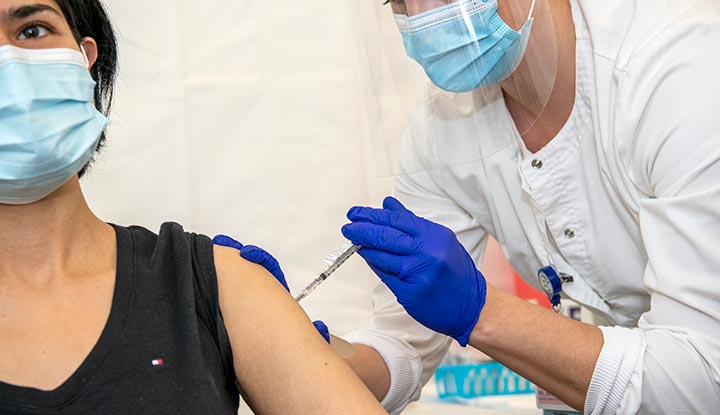 A masked woman is getting an injection in her left arm from a healthcare provider who is wearing gloves, a mask and a face shield.