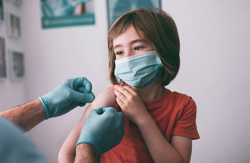 Little girl in mask holds arm while doctor puts on band-aid after vaccine