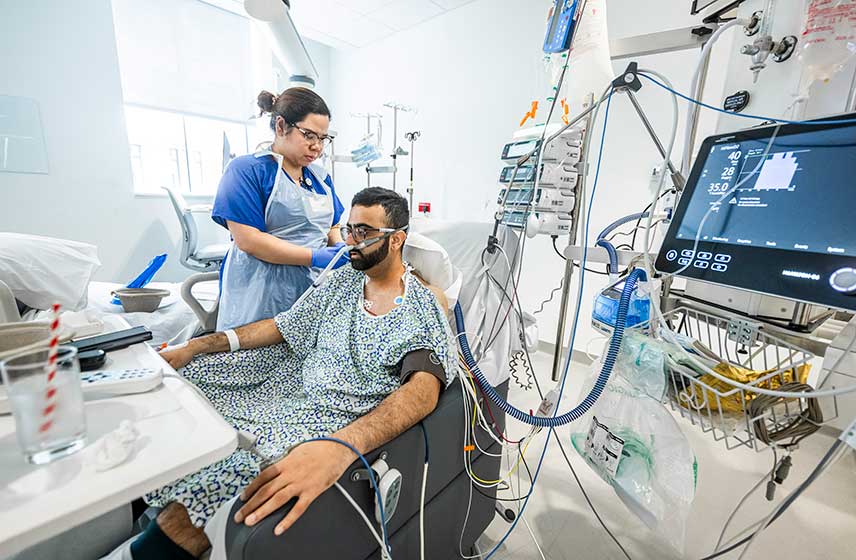 Healthcare worker beside patient hooked to medical devices in hospital room.